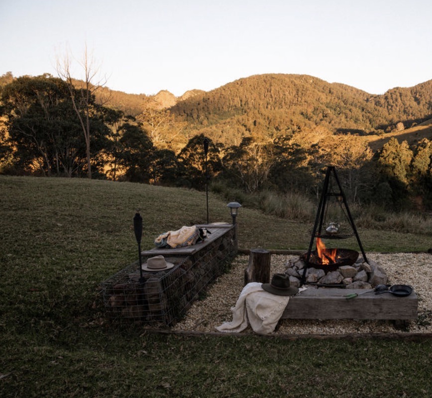A campfire crackling at golden hour with forested mountains glowing behind, at Nature Domes