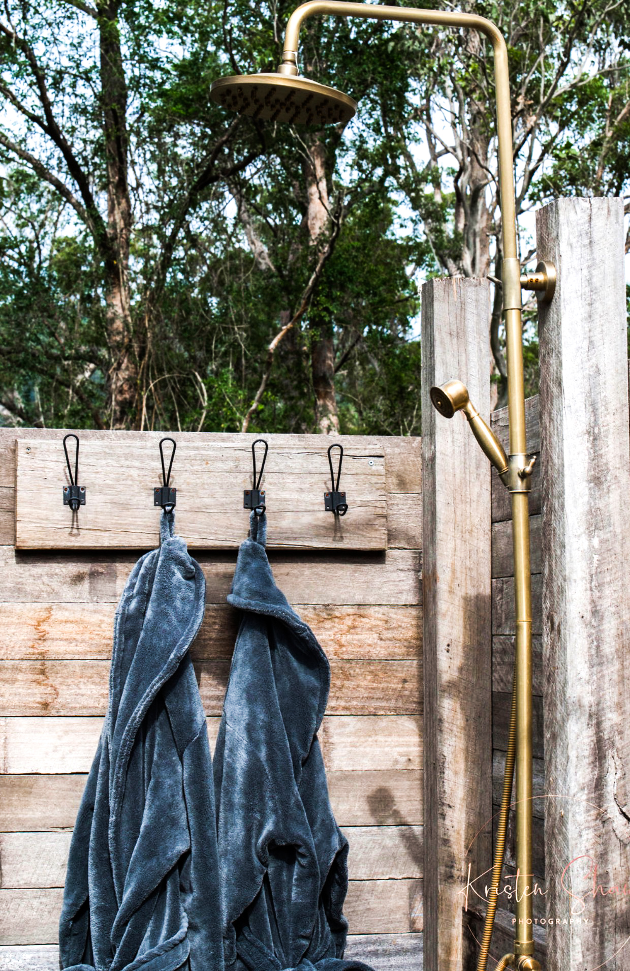 Robes on hooks beside a brass outdoor shower in the trees